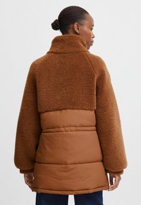 Brun shearling- og polstret jakke med høj krave, der har en struktureret overdel og glat nederste del, brede ærmer og en afslappet pasform.