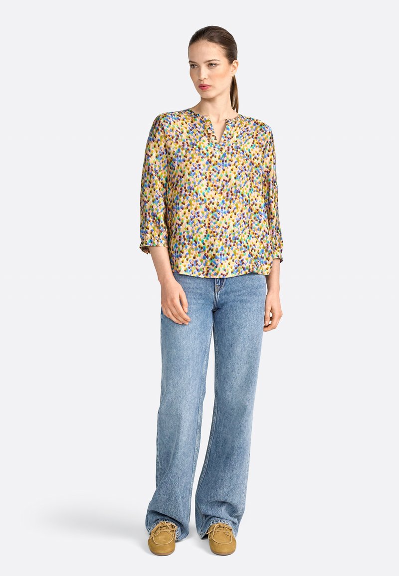 Colorful, patterned blouse with a V-neck and three-quarter sleeves, paired with light blue, loose-fitting jeans and brown suede shoes.