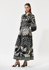 Black and cream patterned dress with long sleeves, cinched waist, and full skirt. Features geometric and floral designs, paired with black heels.