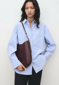 Camicetta azzurro chiaro con tasca frontale, abbinata a una borsa a spalla in pelle marrone scuro, con design curvo.