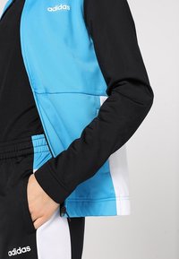 Personne portant une tenue de sport Adidas noire et bleue, main dans la poche, affichant le logo sur la poitrine de la veste et la cuisse du pantalon.