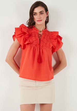 Red ruffled blouse with floral embroidery, tie neckline, and tassel accent. Lightweight fabric, short sleeves, loose fit, paired with a beige skirt.