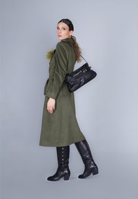Cappotto verde oliva con cintura, caratterizzato da un accento testurizzato. Borsa a spalla nera con dettagli in metallo argentato e stivali neri al ginocchio con dettagli di bottoni.