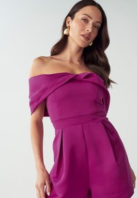 Abito fucsia senza spalline con dettaglio annodato al collo, gonna plissettata e corpino strutturato. Il tessuto sembra liscio e leggero.
