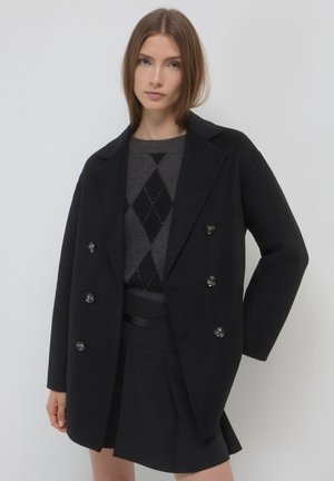 Cappotto di lana nera con colletto a punta, abbottonatura doppiopetto e grandi bottoni. Indossato sopra un maglione scuro a rombi e una gonna nera.