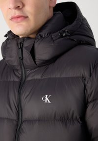 Czarna kurtka puchowa z wysokim kołnierzem i kapturem, z zamkiem błyskawicznym z przodu oraz białym logo "CK" na lewej piersi.