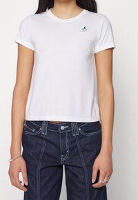 Hvid cropped T-shirt med korte ærmer, der har et lille sort logo-patch og en blød tekstur. Kombineret med mørke denim bukser med hvid syning.