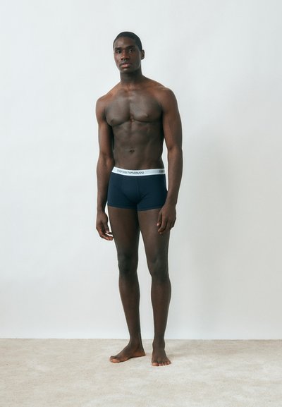 Muskularny mężczyzna stojący boso na tle białej, gładkiej ściany, ubrany w czarne bokserki z białym pasem Emporio Armani.