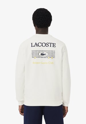 Persona que lleva una sudadera blanca de Lacoste con un diseño de red y pelotas de tenis, y los textos "LACOSTE" y "Roland Garros Club" en la espalda.