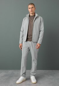 Light gray athletic jacket with a high collar, paired with matching joggers. Features accent stitching, flat seams, and white sneakers.