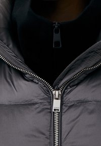 Primo piano di una cerniera di una giacca nera parzialmente aperta sopra un capo in maglia nero con una cerniera più piccola nell'area del colletto.