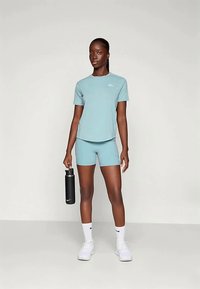 Αθλητική μπλούζα και σορτς γαλάζιου χρώματος από λεία ύφανση. Το μοντέλο κρατάει μια μαύρη φιάλη νερού. Άσπρα αθλητικά παπούτσια και κάλτσες ολοκληρώνουν την εμφάνιση.