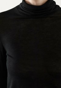 Close-up van een persoon die een zwart coltrui draagt, waarbij de nek, schouders en bovenste gedeelte van de borst zichtbaar zijn.