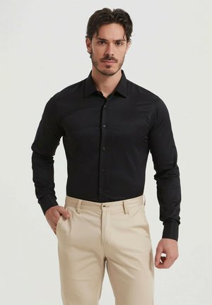Chemise noire à manches longues avec col boutonné, présentant une texture lisse et une coupe standard, associée à un pantalon beige.
