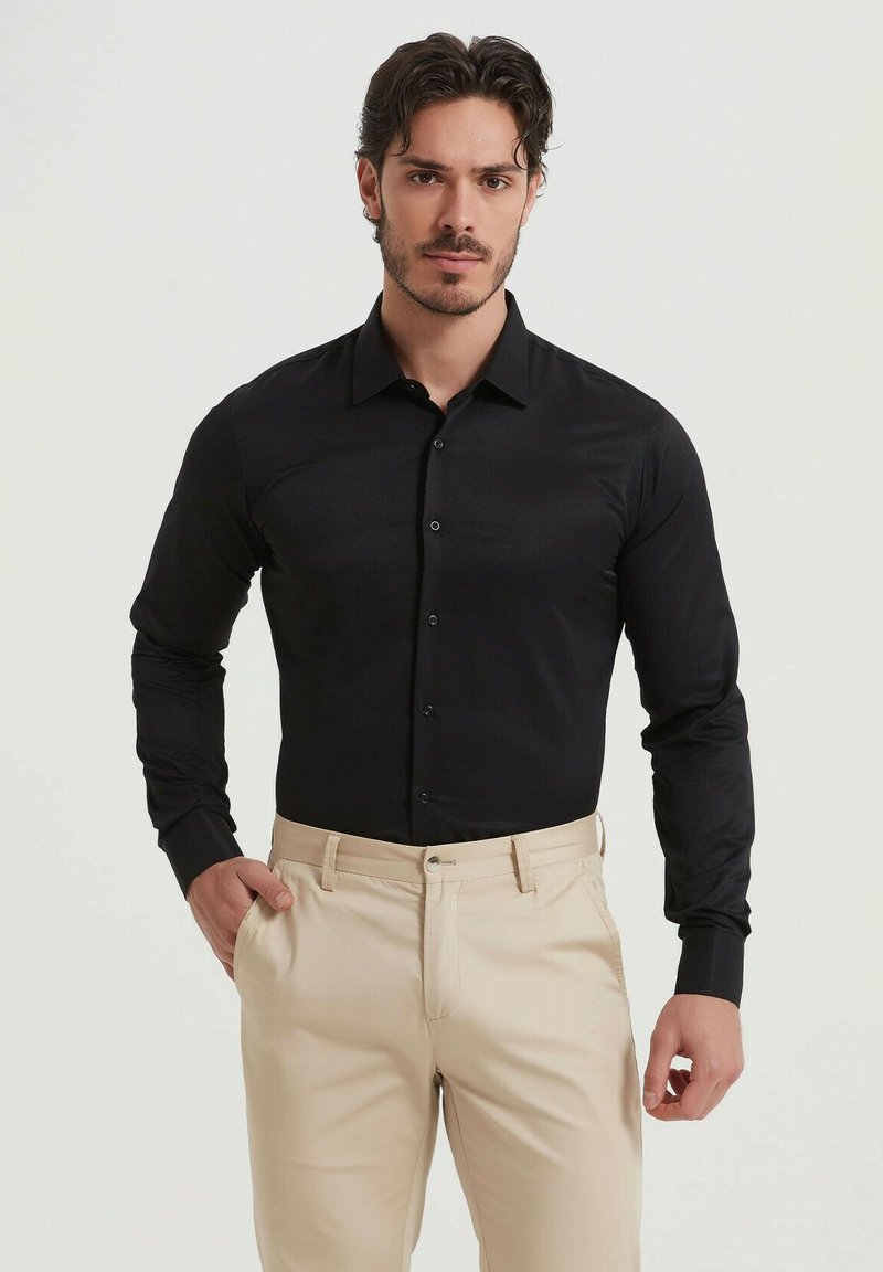 Chemise noire à manches longues avec col boutonné, présentant une texture lisse et une coupe standard, associée à un pantalon beige.