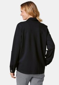 Zwarte langemouwen shirt met een kraag en knoopmanchetten, gemaakt van gladde stof. Draagt een zwart-wit geruit broek met patroon.