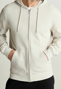 Světlé béžové mikina na zip s texturovanou prodyšnou tkaninou, kapucí se stahovací šňůrkou a předními kapsami; žebrované manžety a lem.