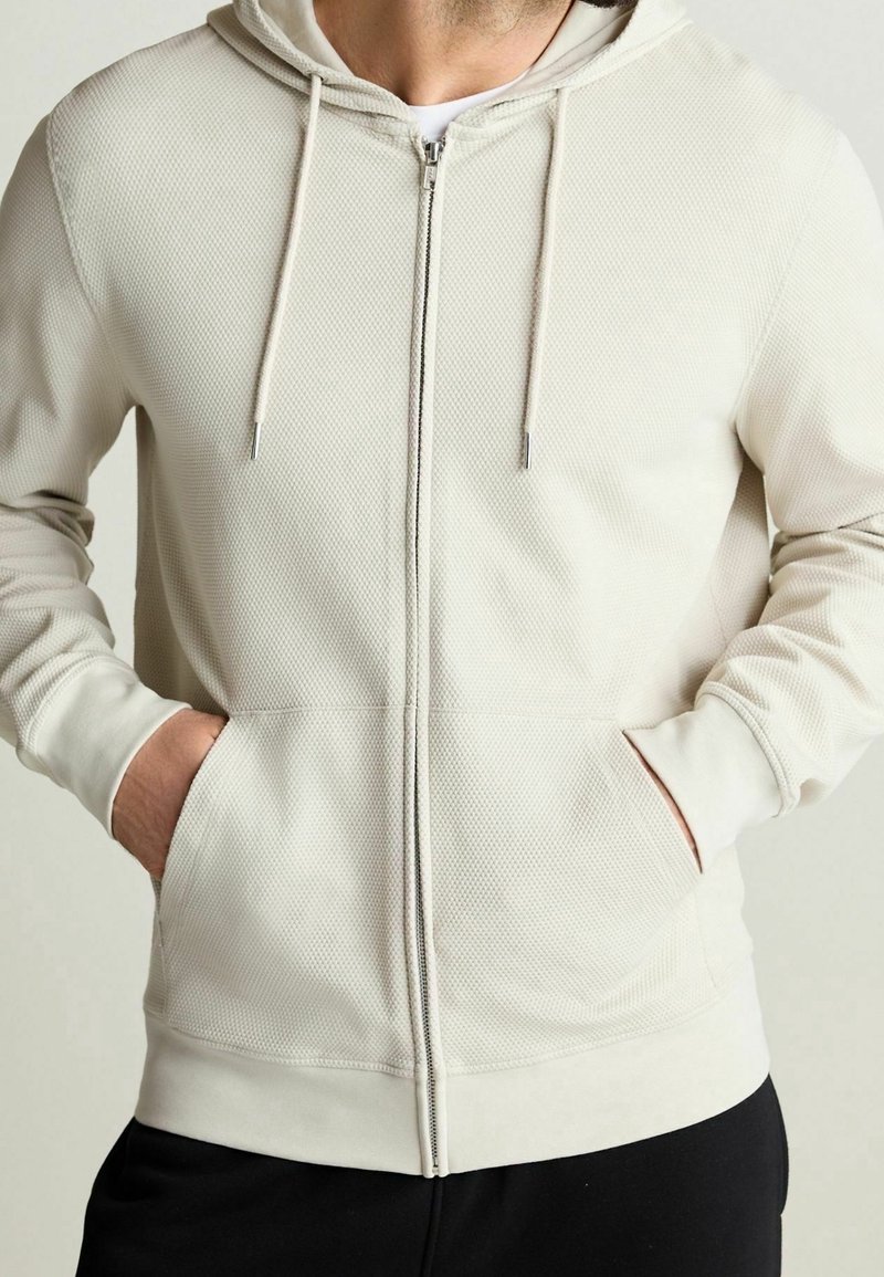 Světlé béžové mikina na zip s texturovanou prodyšnou tkaninou, kapucí se stahovací šňůrkou a předními kapsami; žebrované manžety a lem.