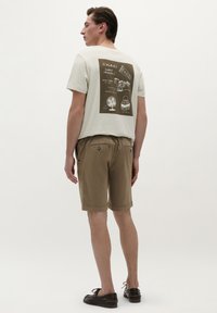 Tricou scurt cu mânecă scurtă de culoare bej deschis, cu grafică ilustrată maro pe spate, asortat cu pantaloni scurți maro și încălțăminte închisă. Design simplu.