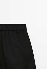 Tissu noir avec une ceinture élastique froncée, probablement une partie d'une jupe ou d'un pantalon, présenté sur un fond blanc uni.