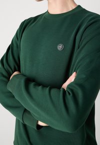 Felpa verde in cotone con collo a giro, caratterizzata da un piccolo logo rotondo nero sul petto e polsini a coste. Texture liscia e design aderente.