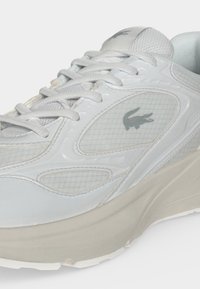 Baskets blanches Lacoste avec panneaux en mesh, lacets et logo sur le côté, avec une semelle épaisse écrue.