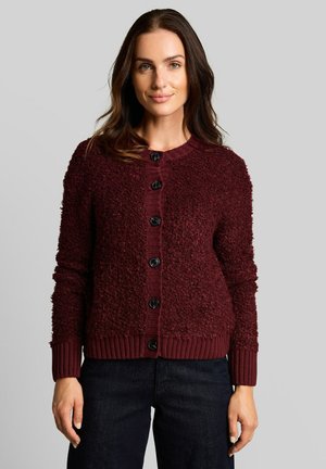 Femme portant un cardigan bordeaux texturé boutonné et un pantalon foncé, debout devant un fond gris clair uni.