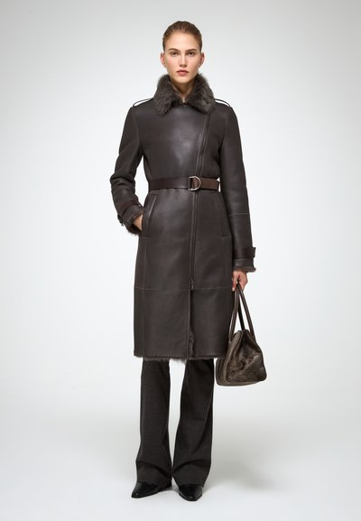 Manteau en cuir marron avec col en fourrure synthétique, taille ceinturée et fermeture éclair à l'avant. Associé à un pantalon sombre et un sac à main texturé.