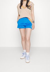 Beige ärmlös tröja med V-ringning och mångfärgade accenter, ihop med ljusblå shorts. Vita sneakers med ljusblå detaljer.