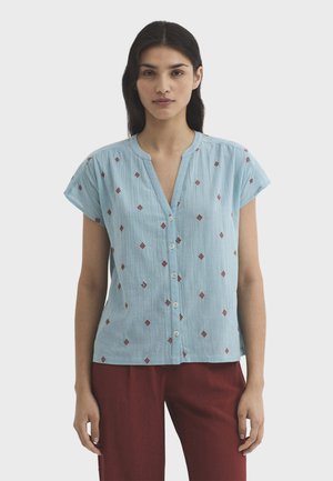 Mujer vestida con una blusa azul claro de manga corta y botones, con pequeños patrones de diamantes rojos, y pantalones burdeos, de pie frente a un fondo liso.