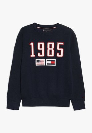 Granatowy sweter z dzianiny z dużym napisem "1985" w białych i czerwonych liczbach, poniżej liczb znajdują się flagi amerykańska i Tommy Hilfiger.