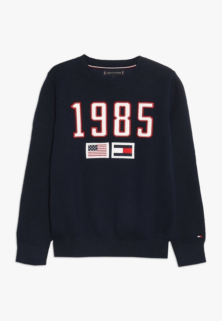 Pull en maille bleu marine avec "1985" en grands chiffres blancs et rouges, comportant les drapeaux américain et Tommy Hilfiger sous les chiffres.