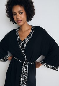 Čierny kimono štýlový župan s trojštvrťovými rukávmi, s čierno-bielym vzorovaným lemom okolo výstrihu a manžiet. Opasok v páse.