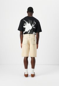 Άνδρας που κοιτάζει προς τα πίσω, φοράει μαύρο μπλουζάκι με γραφικά, μπεζ σορτς μέχρι το γόνατο, λευκές κάλτσες και καφέ δερμάτινα μοκασίνια σε απλό λευκό φόντο.