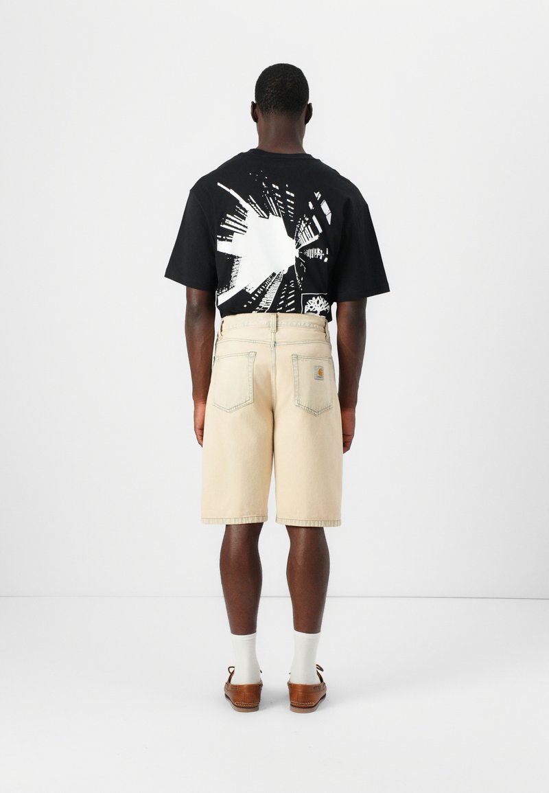 Mand vendt væk iført sort T-shirt med grafik, beige shorts til knæet, hvide sokker og brune læderloafers på ensartet hvid baggrund.