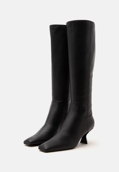Paire de bottes en cuir noires montantes jusqu'aux genoux, avec bouts carrés et talons blocs incurvés uniques, sur un fond clair uni.