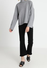 Maglione grigio a collo alto con maniche larghe, abbinato a jeans neri a zampa d'elefante con bordi sfrangiati e scarpe slip-on nere con fibbia.