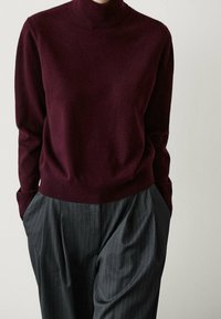 Person in einem bordeauxfarbenen Rollkragenpullover und grauen Streifenanzughosen mit Händen in den Taschen vor einem weißen Hintergrund.