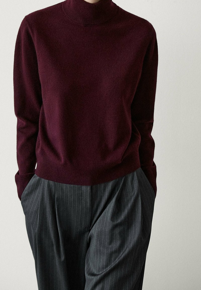 Person in einem bordeauxfarbenen Rollkragenpullover und grauen Streifenanzughosen mit Händen in den Taschen vor einem weißen Hintergrund.