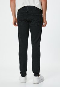 Musta farkkutakki slim-fit-mallilla, jossa on viisi taskua ja ommeltuja yksityiskohtia. Valkoisilla lenkkitossuilla yhdistettynä takaa katsottuna näkyy design.