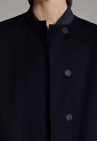Cappotto di lana blu navy con colletto alto e sei bottoni neri. Il tessuto è liscio e leggermente testurizzato, con linee pulite e senza motivi visibili.