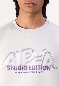 Jasnoszara bluza z dużym, błyszczącym, wielokolorowym logo "ALPHA" z efektem 3D oraz napisem "STUDIO EDITION" w fioletowym kolorze poniżej.