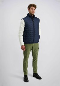 Marineblauwe gewatteerde vest met een hoge kraag, voorzien van een ritssluiting aan de voorkant en twee zijzakken. Gecombineerd met lichtgroene cargobroeken en zwarte schoenen.