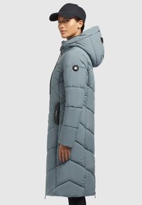Padded dlhý kabát svetlo modrej farby s kapucňou, ktorý má zips a bočné vrecká. Dizajn obsahuje geometrické prešívané vzory.