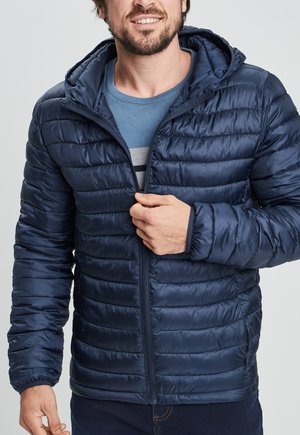 Giacca puffa blu navy con chiusura zip, cappuccio e polsini a coste. Presenta un motivo trapuntato e una finitura lucida per l'isolamento.
