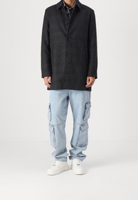 Hombre vistiendo un abrigo abotonado de cuadros gris oscuro, pantalones cargo vaqueros azul claro con múltiples bolsillos y zapatillas blancas, de pie sobre un fondo blanco.