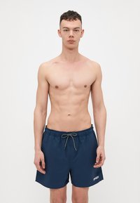 Mørkeblå svømmeshorts med snor i taljen, laget av hurtig tørkende stoff, med en hvit logo på venstre kant.