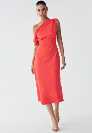 Frau in einem rot, eintägigen, knielangen Kleid mit tailliertem Oberteil und fließendem Rock, die selbstbewusst vor einem neutralen Hintergrund steht.