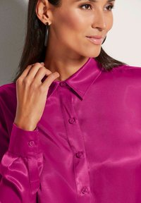 Fuchsia Satinbluse mit strukturiertem Kragen, durchgehender Knopfleiste und umgeschlagenen Ärmeln, die einen subtilen Glanz und eine glatte Textur aufweist.