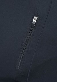 Tessuto nero con una tasca verticale con zip nera, caratterizzata da un tirante della zip in argento e nero e cuciture visibili.